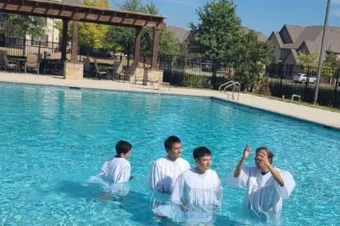 Four people in white baptismal robes standing waist-deep in an outdoor pool, one of them raising a hand in prayer