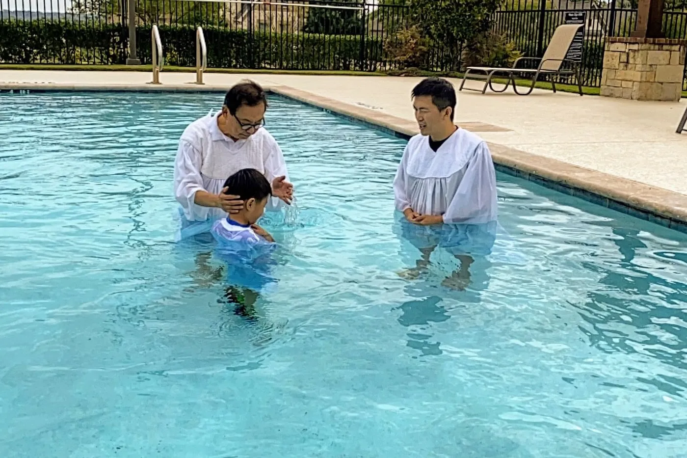 A boy being baptized in an outdoor pool, one church leader resting a hand on his head while another stands alongside in a white robe