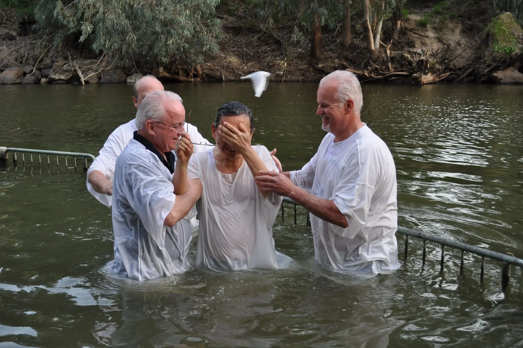 A woman being baptized in a river, held steady by two others in white robes as she comes up out of the water