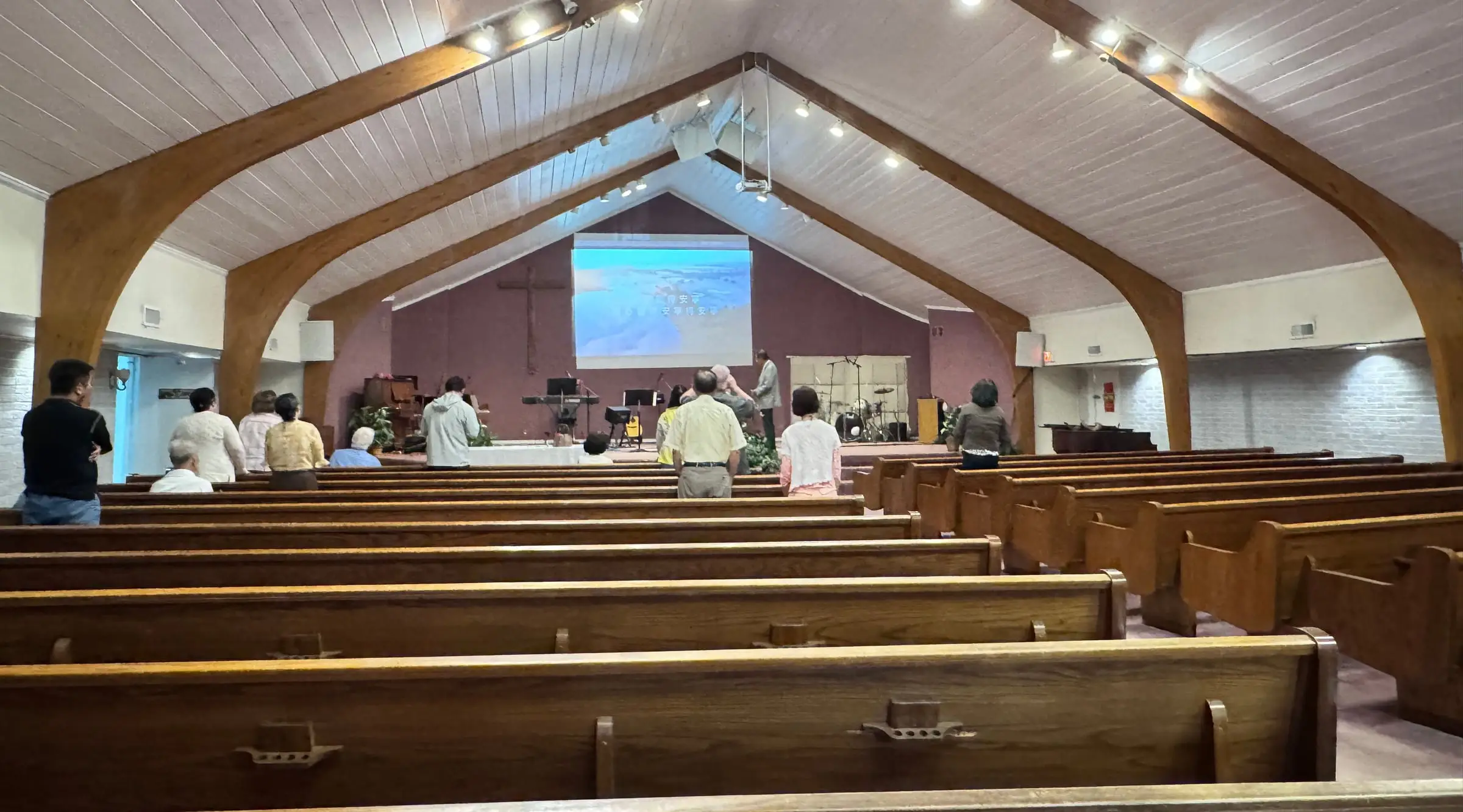 The sanctuary seen from the back row: wooden pews under a white vaulted ceiling with exposed timber arches, the congregation standing at the front beneath a wooden cross