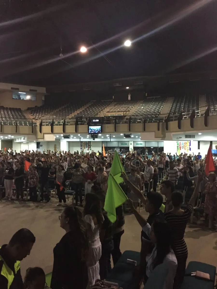 A wide view of a packed arena, hundreds standing with hands raised and coloured flags held up above the crowd