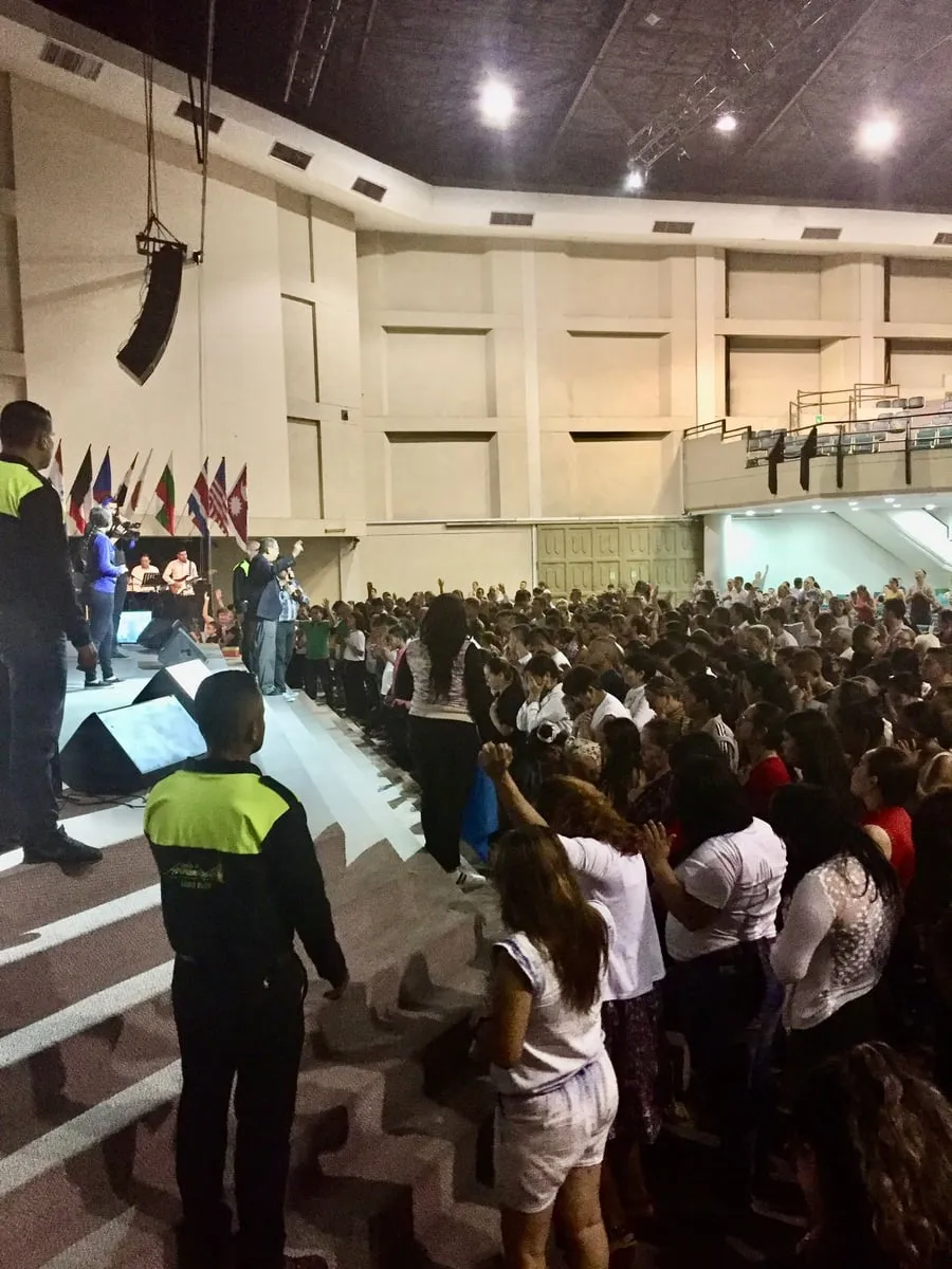 The congregation standing and responding during a conference, many with hands raised, seen from the side of the stage
