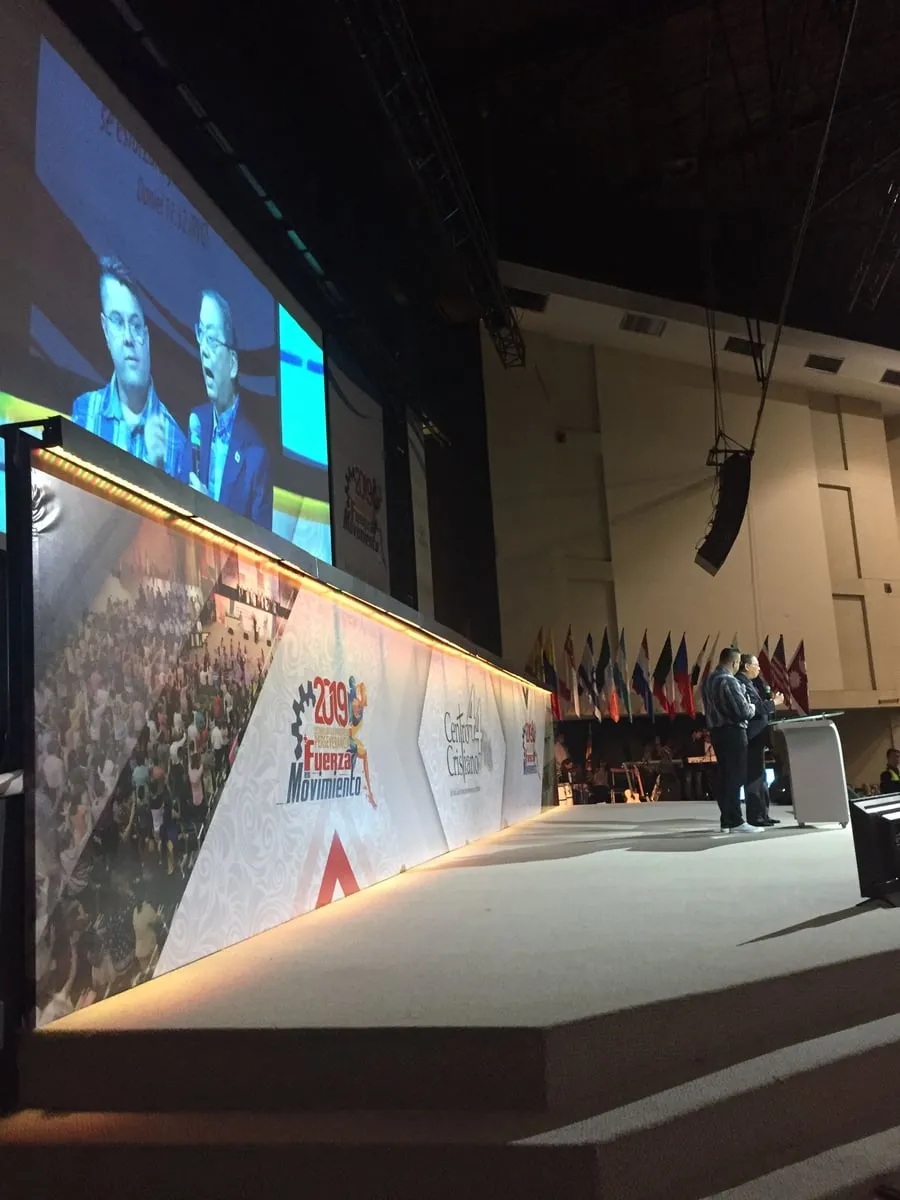 A conference auditorium with Pastor Daniel S. Chen preaching on the large screens beside a translator, rows of national flags lining the stage behind him