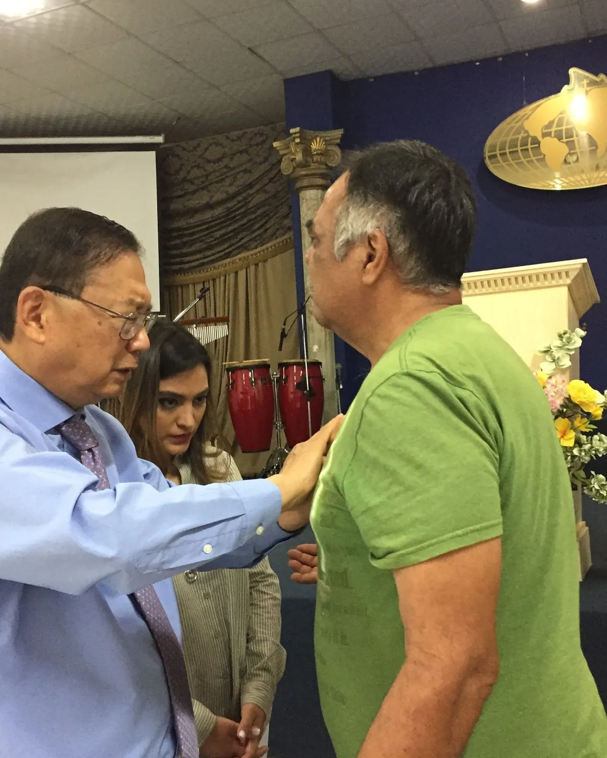 Pastor Daniel S. Chen praying with a man, one hand resting on his shoulder, while another member of the church stands alongside